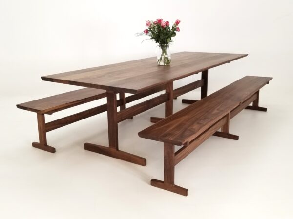 A BRIT Trestle Dining Table with a beveled edge, two benches, and a vase of flowers.