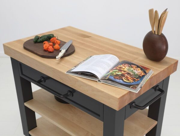An close up image of a butcher block kitchen cart with a cook book and vegetables on top.